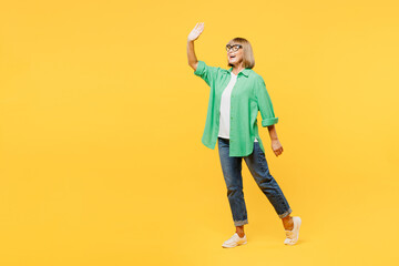 Fototapeta na wymiar Full body elderly side profile view smiling blonde woman 50s years old wear green shirt glasses casual clothes walking going waving hand isolated on plain yellow background studio. Lifestyle concept