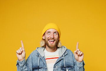 Young smiling happy fun blond man he wear denim shirt hoody beanie hat casual clothes point index finger overhead on area mockup isolated on plain yellow background studio portrait. Lifestyle concept