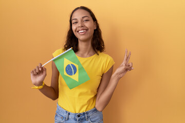 Young hispanic woman holding brazil flag smiling with happy face winking at the camera doing...