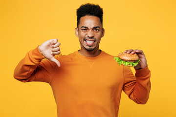 Young displeased man wear orange sweatshirt casual clothes eat burger show thumb down gesture...