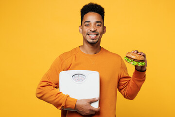 Young man of African American ethnicity wear orange sweatshirt casual clothes eat burger hold...