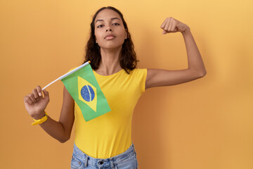 Young hispanic woman holding brazil flag strong person showing arm muscle, confident and proud of power