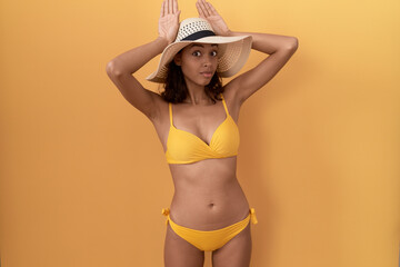 Young hispanic woman wearing bikini and summer hat doing bunny ears gesture with hands palms looking cynical and skeptical. easter rabbit concept.