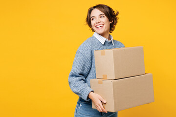 Young smiling cheerful woman wears grey knitted sweater shirt casual clothes hold cardboard boxes look aside on area workspace isolated on plain yellow background studio portrait. Lifestyle concept.