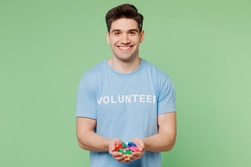 Young happy man wear blue t-shirt white title volunteer hold in hand plastic bottle cups to recycle isolated on plain pastel green background Voluntary free work assistance help charity grace concept