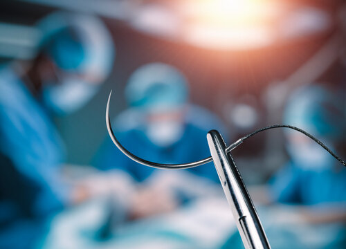 Hemostatic forceps holding half circle cutting needle and suture in front of of blurry operators under the lights in operating room.