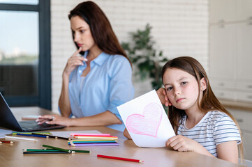 Bored child holding greeting card with heart to busy mother.