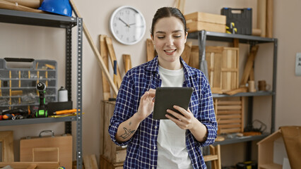 A young woman in a checkered shirt uses a tablet in a well-organized carpentry workshop.
