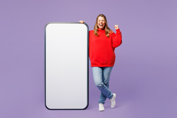 Full body young woman wearing red hoody casual clothes big huge blank screen mobile cell phone smartphone with workspace copy space mockup area do winner gesture isolated on plain purple background.