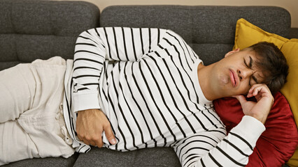 Portrait of a worried young hispanic man suffering from agonizing stomachache, lying on the sofa in...