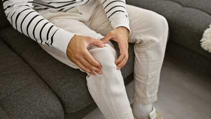 Hispanic man's hands gripping knee in pain, sitting on comfy living room sofa, suffering from leg...