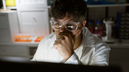 Handsome, serious-faced young hispanic male scientist engrossed in high-stakes research, working diligently with computer in dim laboratory, amidst glowing test tubes and microscope.