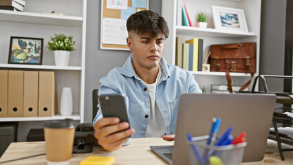 Handsome young hispanic man, a successful business professional, boss and manager, at work intensely using his smartphone and laptop, seriously concentrated yet relaxed in elegant office background. - obrazy, fototapety, plakaty