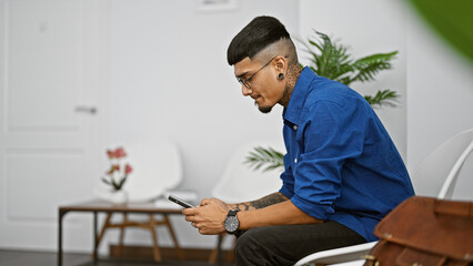 Handsome young latin man engrossed in texting on smartphone while sitting in a busy room, waiting...