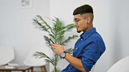Handsome young latin man, tattooed and serious, engrossed in his smartphone while sitting in a...
