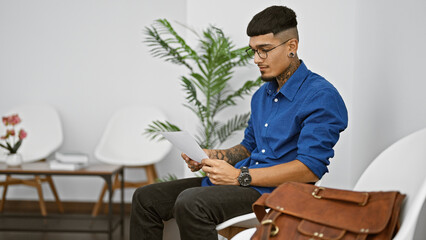 Handsome latin man, a portrait of a young, tattooed professional, doubting his ideas as he concentrates on reading a document, while sitting in a waiting room chair, pensive