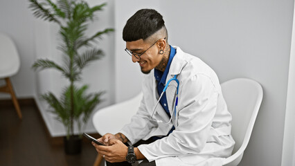 Happy young latin man, a confident doctor, smiling while using his smartphone, sits in the clinic's waiting room, fully engrossed in his professional work, portraying a portrait of modern healthcare.