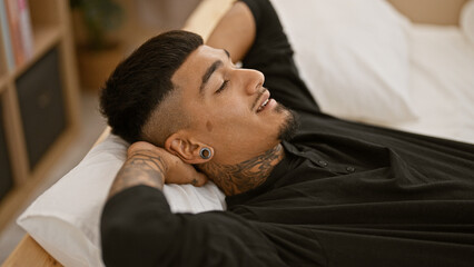 Handsome, tattooed young latin man lying on bed, hands resting on head, radiating confidence and relaxation as he rouses from sleep, a portrait of carefree indoor lifestyle