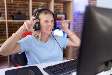 Young caucasian woman playing video games wearing headphones smiling funny doing claw gesture as...