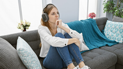 Pensive young caucasian woman wearing headphones sitting on a gray sofa in a cozy living room.