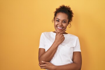 Young hispanic woman with curly hair standing over yellow background looking confident at the camera smiling with crossed arms and hand raised on chin. thinking positive.