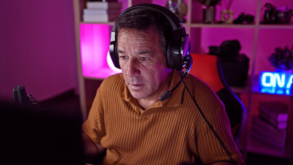 Mature man with headphones focused on computer in vibrant gaming room.