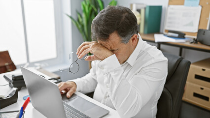 A stressed businessman in an office removing his glasses in frustration.