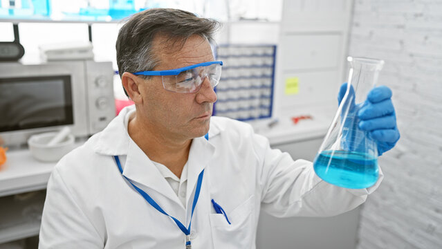 A Mature Man Scientist Examines A Blue Liquid In A Flask In A Laboratory Setting Wearing Protective Eyewear.