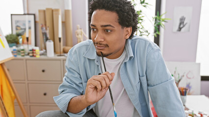 A thoughtful young man with curly hair holds a paintbrush in a bright, artistic interior studio.