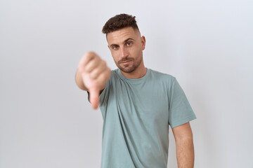 Hispanic man with beard standing over white background looking unhappy and angry showing rejection and negative with thumbs down gesture. bad expression.