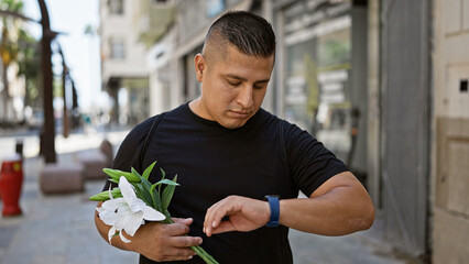 Cool young latin man standing on a sunny city street, looking at his watch, hand grasping a bouquet...