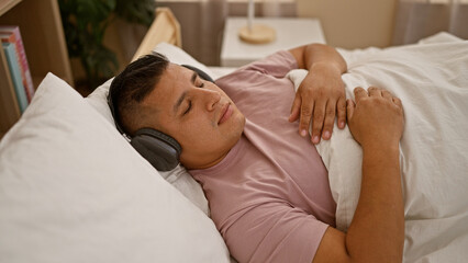 Cozy morning vibes, young latin man comfortably lying in bed, relaxing and wearing headphones,...