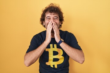Hispanic young man wearing bitcoin t shirt laughing and embarrassed giggle covering mouth with...