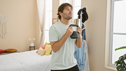 A focused young caucasian man lifts a kettlebell in his bright bedroom, exemplifying home fitness.