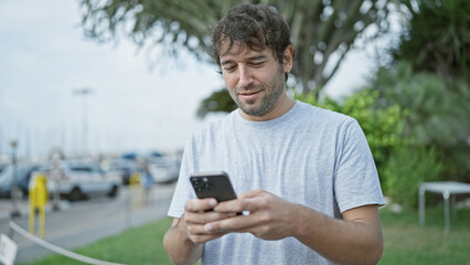 Happy young man, sporting a beard and blond hair, enjoying his time outdoor in the city, confidently smiling while typing a message on his smartphone screen