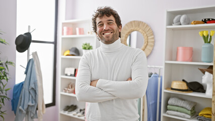 Handsome hispanic man with beard smiling in a modern wardrobe room, arms crossed, wearing a white...