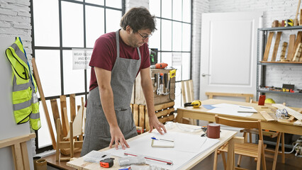 A focused hispanic man reviews plans in a sunny carpentry workshop, representing craftsmanship and...
