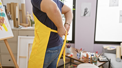 A man in a yellow apron ties the straps in an art studio with paintings and brushes visible.