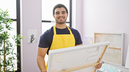 A smiling man with a beard, wearing an apron, holds a canvas in a bright art studio.