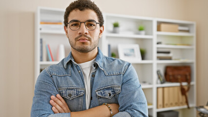 Casual young man with glasses and beard standing arms crossed in a modern office setting.