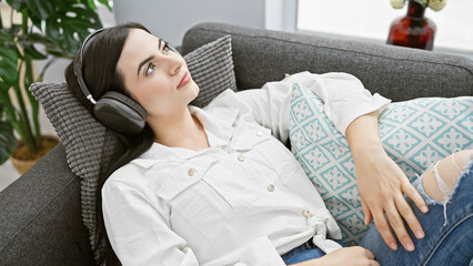 A relaxed young woman enjoys music on headphones while reclining on a sofa, creating a serene...