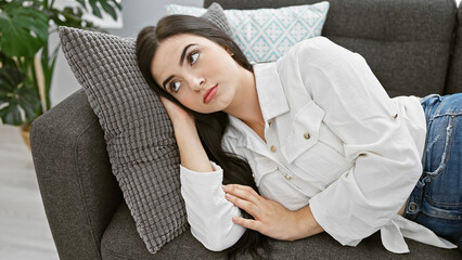 Pensive young hispanic woman lounging on a sofa indoors, projecting beauty and casual elegance in a comfortable home setting.