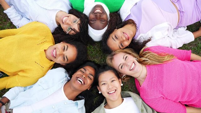 Multiracial group of young women in circle smiling at camera together - Happy girlfriends having fun lying on the grass at the park - Female community and friendship concept - 4k video