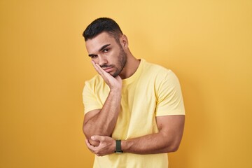 Young hispanic man standing over yellow background thinking looking tired and bored with depression...