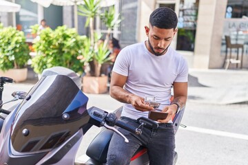 Young latin man counting dollars sitting on motorbike at street