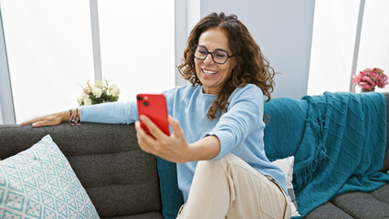 A smiling middle-aged hispanic woman with curly hair takes a selfie indoors on a couch, depicting a...