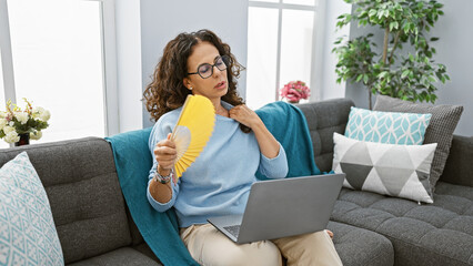 Mature hispanic woman with curly hair using fan while working on laptop in a cozy living room.