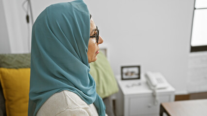 A mature muslim woman with glasses and a hijab looks pensive in a cozy living room setting.