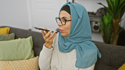 A mature woman wearing glasses and a hijab speaks into a smartphone in a cozy living room.