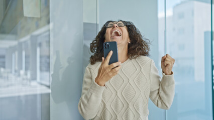 Fototapeta premium Excited hispanic woman celebrating with smartphone in modern office setting